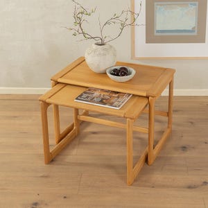 Può includere: Due tavolini ad incastro in legno chiaro. Il tavolo superiore contiene una piccola ciotola bianca con frutta scura e una rivista. Un vaso decorativo con rami è posto sopra. I tavoli sono su un pavimento in legno chiaro.