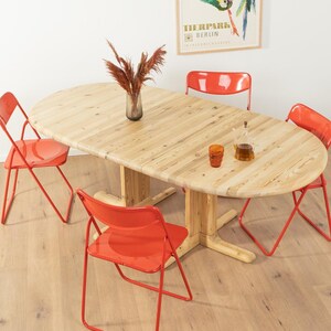 Peut inclure: Une table à manger ovale en bois clair avec un pied central, entourée de quatre chaises en métal rouge. Un vase avec des fleurs séchées et quelques objets en verre sont sur la table. La table mesure environ 152 cm de long.