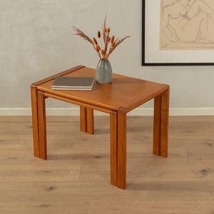 Op de afbeelding: Een vierkante houten bijzettafel met twee niveaus en een warme, roodbruine afwerking. Een kleine, gestreepte vaas met gedroogde bloemen en een boek rusten op de bovenkant. De tafel staat op een parketvloer.