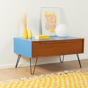 Op de afbeelding: Een retro-stijl kast met een lichtblauw blad en een houten lade. De kast heeft zwarte haarspeldpoten. Decoratieve items zijn gele vazen, boeken, een witte bol en een ingelijst abstract kunstwerk.