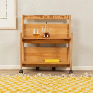 May include: A light-colored wooden serving cart with two shelves and small wheels. The top shelf holds a glass of liquid and a glass carafe. A yellow book is on the bottom shelf. The cart is on a patterned rug.