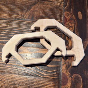 May include: Two light-colored wooden frames with an octagonal inner cutout and angled outer edges. Each frame features two distinct notched hook-like protrusions on the sides, crafted from smooth, unfinished plywood with visible wood grain.