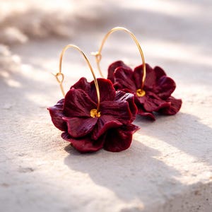 Könnte beinhalten: Burgunderfarbene Blumenohrringe mit goldfarbenen Reifen. Die Ohrringe haben geschichtete Blütenblätter und eine kleine goldene Mitte. Die Ohrringe werden auf einer hellen Oberfläche präsentiert.
