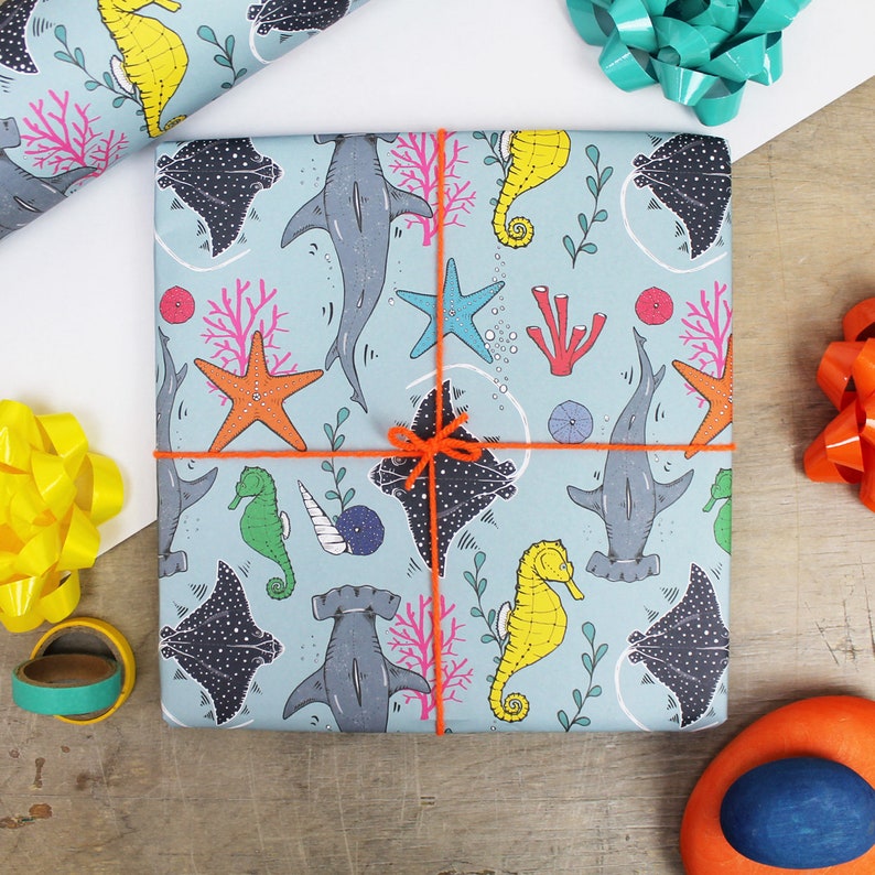 May include: A wrapped gift with blue wrapping paper featuring a sea life pattern of hammerhead sharks, seahorses, stingrays, and starfish. The gift is tied with orange twine and surrounded by colorful bows.