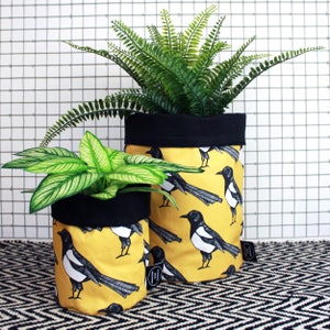 Peut inclure: Deux pots de plantes en tissu avec une bordure noire, présentant un motif de pie noir et blanc sur fond jaune. Le plus grand pot contient une plante de fougère verte, et le plus petit pot contient une plante verte feuillue.