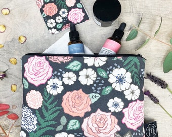 Op de afbeelding: Een zwarte toilettas met een bloemenprint op een witte achtergrond. De tas is gevuld met twee kleine glazen flesjes met etiketten waarop "Faceberry" en "Centella Asiatica Botanical Serum" staat. De tas ligt op een houten oppervlak met een witte washandje en een klein potje.