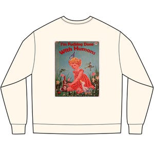 Può includere: Felpa color crema con una grafica di una copertina di libro vintage. L'illustrazione della copertina mostra una bambina in un vestito rosso con un coniglio e un'anatra, con il testo "I'm Fucking Done With Humans".