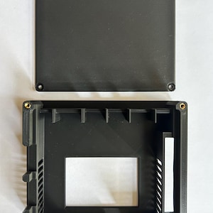 May include: Black electronic device enclosure with a removable top. The top has a textured surface and is secured with four screws. The bottom section has an open interior with structural supports and a rectangular opening.