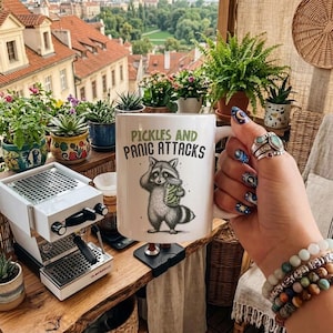 Può includere: Tazza in ceramica bianca con la scritta "Pickles and Panic Attacks" e un'illustrazione di procione. La tazza è tenuta davanti a un balcone con piante e vista sulla città.