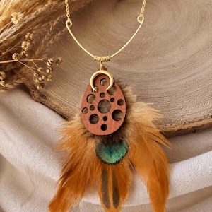 Peut inclure: Un collier doré composé d'une fine chaîne et d'une barre métallique incurvée. Le pendentif est une pièce en bois brun rougeâtre circulaire avec des trous découpés, surmontée d'un petit croissant de lune doré et ornée de plumes brunes.