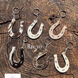 Peut inclure: Six porte-clés en forme de fer à cheval avec différents motifs de poils de vache dans des tons marron, blanc, noir et beige. Chaque porte-clés comprend un anneau et une chaîne en métal argenté. Le mot RECTO est imprimé en blanc au centre.