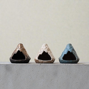 Könnte beinhalten: Drei keramische, bergförmige Dekorationsstücke in Bronze, gesprenkeltem Beige und Blau. Jedes hat eine dunkle Öffnung an der Basis, die einer Höhle ähnelt. Die Objekte sind auf einer grauen Oberfläche angeordnet.