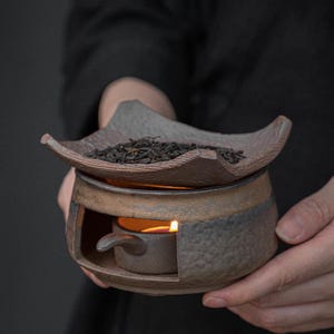May include: A handcrafted ceramic tea warmer with a candle inside. The tea warmer has a dark brown and bronze color scheme, with a textured surface. A small amount of loose tea sits on top of the warmer.