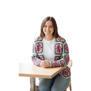 Op de afbeelding: Een kleurrijk gehaakt vest met een bloemenpatroon in paars, roze, groen en crème. De open trui wordt over een wit t-shirt gedragen. De persoon zit aan een lichtgekleurde houten tafel.