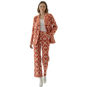May include: A two-piece suit in orange with a cream geometric pattern. The blazer has a classic lapel and the trousers are wide-legged. The model is wearing a white tank top and white boots. The suit is a striking ensemble, suitable for various occasions.