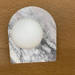May include: A white, spherical object rests on a marble base with gray and white veining. The base has an arched shape. The background is a solid brown color.