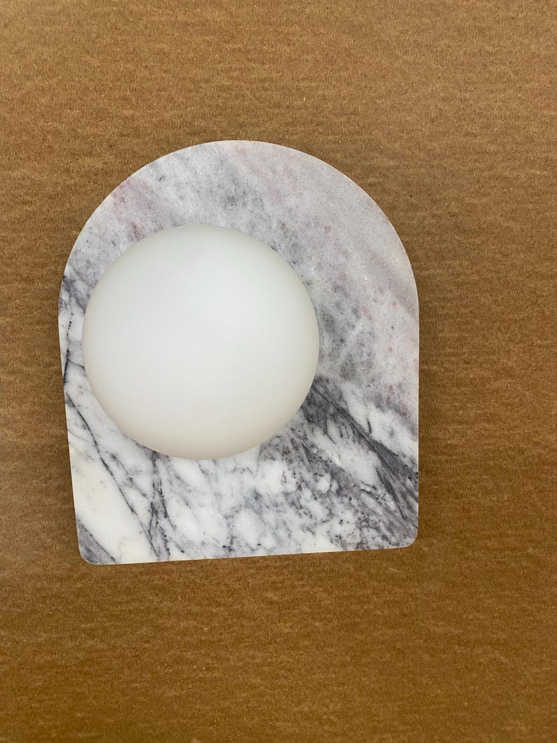 May include: A white, spherical object rests on a marble base with gray and white veining. The base has an arched shape. The background is a solid brown color.