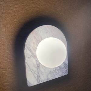 May include: A modern wall sconce featuring a white, spherical light source set against a marble arch. The marble has gray and pink veining. The sconce is mounted on a brown surface.