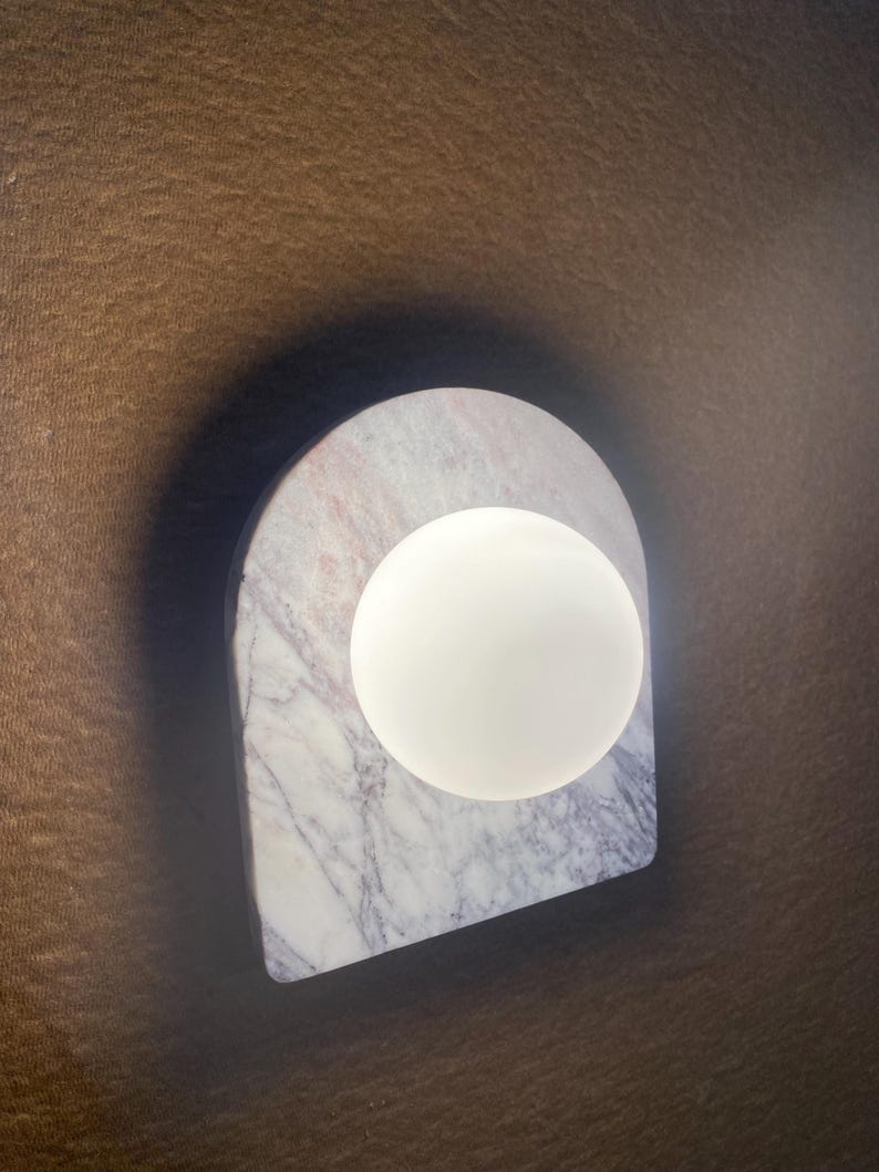 May include: A modern wall sconce featuring a white, spherical light source set against a marble arch. The marble has gray and pink veining. The sconce is mounted on a brown surface.