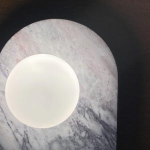 May include: A modern lamp featuring a white, spherical light source set against a marble base. The marble has a gray and pink veined pattern, with an arched shape. The background is dark.