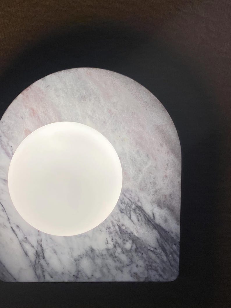 May include: A modern lamp featuring a white, spherical light source set against a marble base. The marble has a gray and pink veined pattern, with an arched shape. The background is dark.