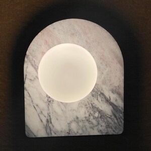 May include: A modern lamp featuring a white, spherical light source set within a marble arch. The marble has a gray and pink veined pattern. The lamp is designed with a minimalist aesthetic.