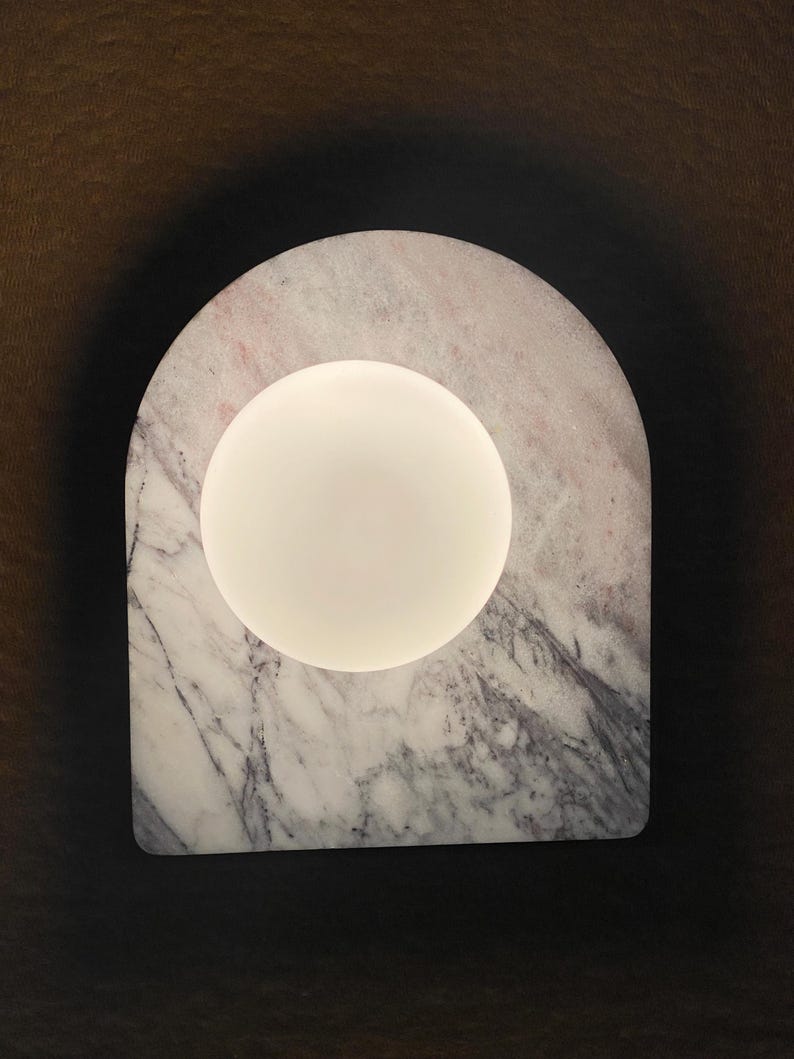 May include: A modern lamp featuring a white, spherical light source set within a marble arch. The marble has a gray and pink veined pattern. The lamp is designed with a minimalist aesthetic.