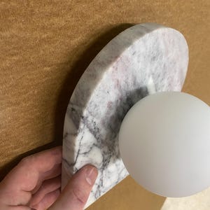 May include: A modern wall sconce featuring a semi-circular marble base with gray and pink veining, paired with a white spherical light fixture. The sconce is held against a brown background.