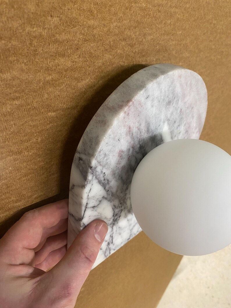 May include: A modern wall sconce featuring a semi-circular marble base with gray and pink veining, paired with a white spherical light fixture. The sconce is held against a brown background.
