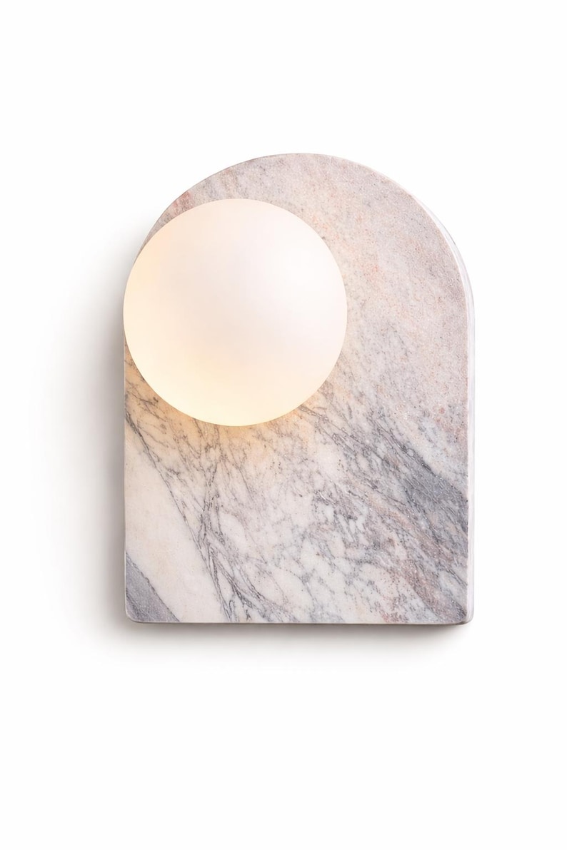 May include: A modern wall sconce featuring a marble base with a rounded top and a frosted glass globe. The marble has a gray and pink veining pattern. The globe is lit, emitting a soft glow.