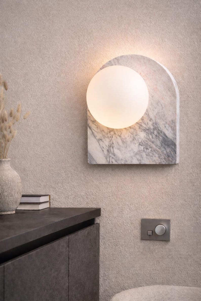 May include: A modern wall sconce featuring a marble backplate with gray veining and an arched top. A round, frosted glass globe provides soft illumination. The sconce is mounted on a textured beige wall.
