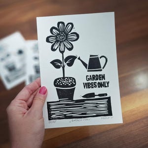 May include: Black and white linocut print featuring a large flower in a pot, a watering can, and a trowel. The text "GARDEN VIBES ONLY" is printed below the watering can. The print is on white paper.