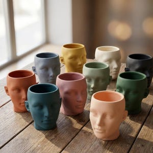 Może przedstawiać: Kolekcja ceramicznych donic w kształcie głowy w różnych kolorach, w tym pomarańczowym, niebieskim, różowym, żółtym, zielonym i czarnym. Donice są ustawione na drewnianej powierzchni, prezentując szczegółowe rysy twarzy i otwarte górne części na rośliny.