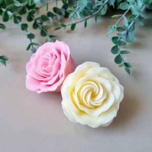May include: Two rose-shaped soaps. One is light pink, the other is pale yellow. The petals are intricately detailed, creating a realistic floral appearance. Green foliage is in the background, adding a natural touch. The soaps are on a neutral-toned surface.