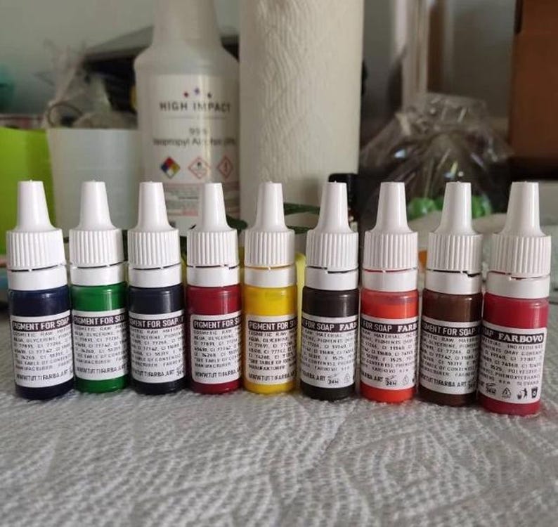 May include: A row of small, colorful pigment bottles for soap making. The bottles are white with white caps and contain various colors, including blue, green, red, and yellow. The labels read "PIGMENT FOR SOAP".