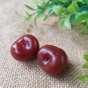 May include: Two dark red, glossy cherries with a small indentation at the stem end. The cherries are set against a textured, tan background with green foliage in the upper right corner.