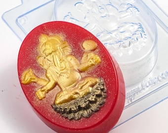 May include: A red, oval-shaped soap with a gold-colored Ganesh design. The soap has a glittery finish and is next to a clear plastic mold with the same Ganesh design. The soap is a handmade item.