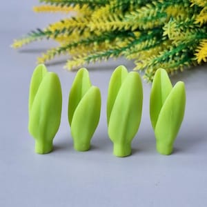 Può includere: Quattro gomme a forma di tulipano, di colore verde chiaro, sono disposte in fila. Le gomme hanno una consistenza liscia e una base arrotondata. Sullo sfondo, una composizione sfocata di fiori artificiali gialli e verdi.