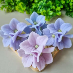 Op de afbeelding: Close-up van verschillende kunstmatige hortensia bloemen in tinten lavendel, lichtblauw en roze. De bloemen zijn gerangschikt op een kleine, decoratieve houten basis, met groen gebladerte op de achtergrond.