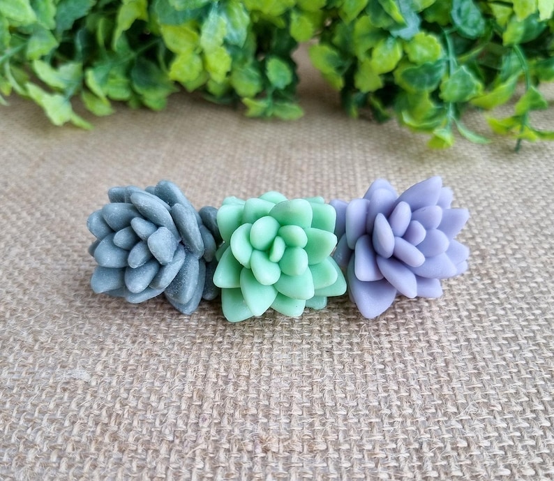 May include: Three succulent-shaped soaps in shades of gray, green, and lavender. The soaps are arranged in a row, showcasing their detailed petal formations. The background features a textured, neutral surface and greenery.