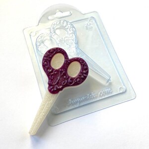 May include: A purple and white scissors-shaped sweet on a clear plastic mould. The scissors have a decorative handle design and a glittery white blade. The mould is also in the shape of scissors.