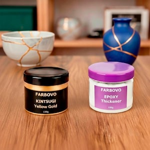 May include: Two FARBOVO Kintsugi product jars: a black jar with yellow gold trim and a white jar with a purple lid. The black jar reads "Kintsugi Yellow Gold 150g". The white jar reads "Epoxy Thickener 150g". A white bowl and blue vase are in the background.