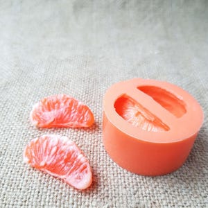 May include: Orange-colored soap mold in the shape of a citrus fruit slice, with two soap slices next to it. The soap slices are a vibrant orange with white accents, resembling a grapefruit or orange segment. The mold is round and has two cavities.