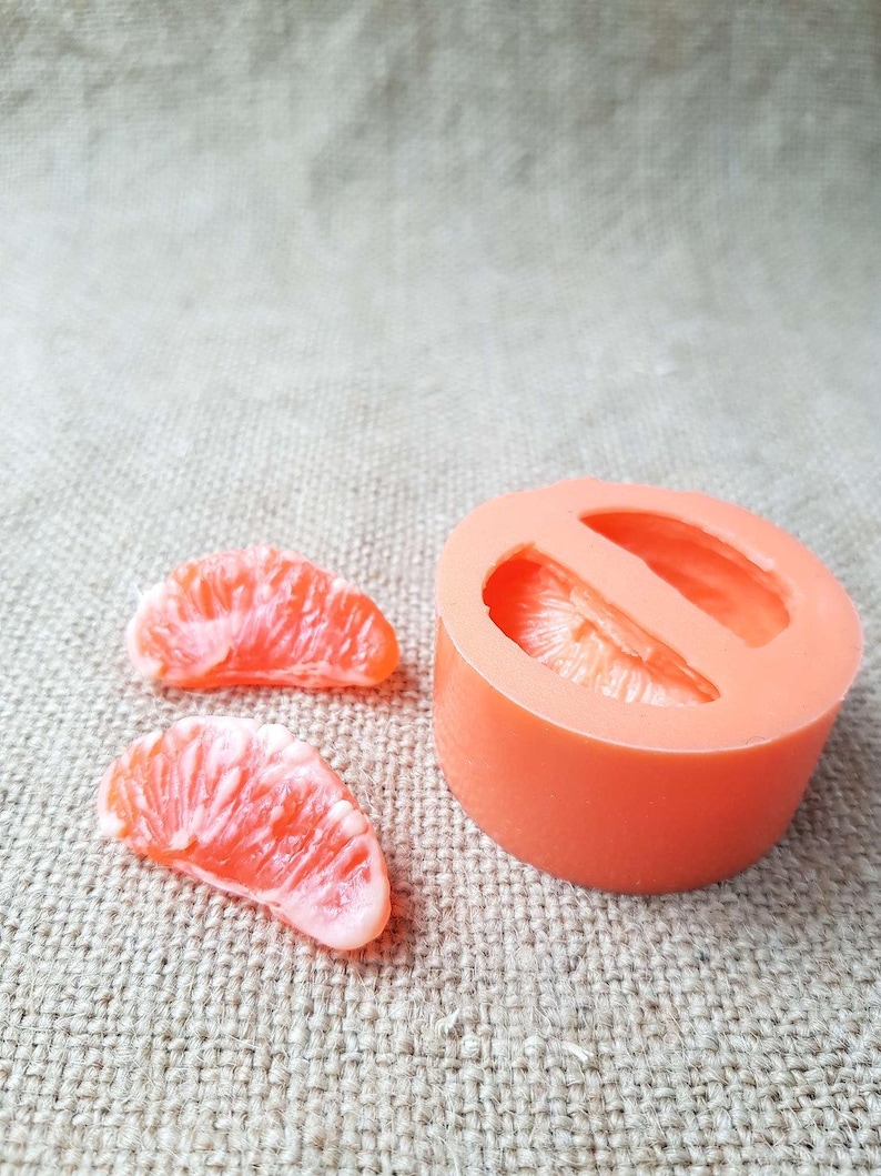 May include: Orange-colored soap mold in the shape of a citrus fruit slice, with two soap slices next to it. The soap slices are a vibrant orange with white accents, resembling a grapefruit or orange segment. The mold is round and has two cavities.