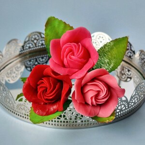 May include: Three artificial roses in shades of red and pink sit on a decorative silver tray. The roses have green leaves and are arranged to showcase their intricate petal details. The tray has a delicate, openwork design.