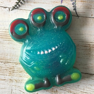 May include: A teal-colored, glittery soap shaped like a cartoon alien with three eyes. The eyes have red and black outlines with yellow centers. The alien has a smiling mouth with white teeth and gray feet with yellow accents.