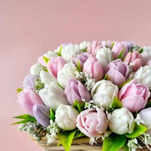 Pode incluir: Uma cesta transbordando com tulipas brancas, rosas e lilás. Folhas verdes e pequenos detalhes brancos são intercalados entre as flores. O fundo é rosa claro liso.
