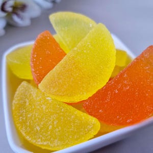 May include: A close-up of a white dish filled with colorful, fruit-shaped candies. The candies are in shades of yellow and orange, with a textured, sugared surface. The candies are arranged in a close-up shot, with a blurred background.