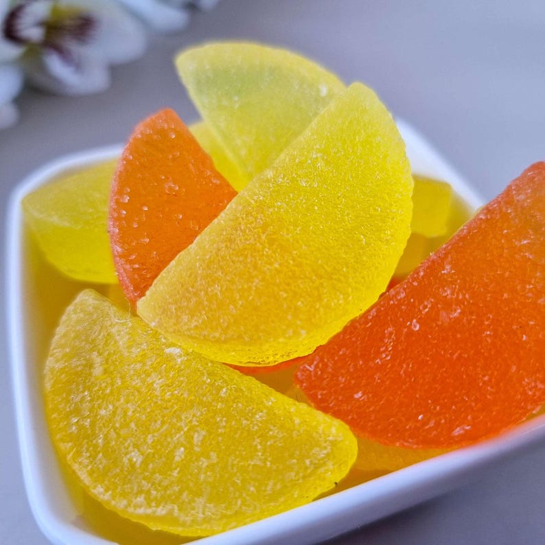 May include: A close-up of a white dish filled with colorful, fruit-shaped candies. The candies are in shades of yellow and orange, with a textured, sugared surface. The candies are arranged in a close-up shot, with a blurred background.