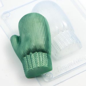 May include: A green mitten-shaped mould with a textured surface, next to a clear plastic mould of the same shape. The mitten mould has a ribbed cuff and a detailed pattern. The clear mould is transparent.